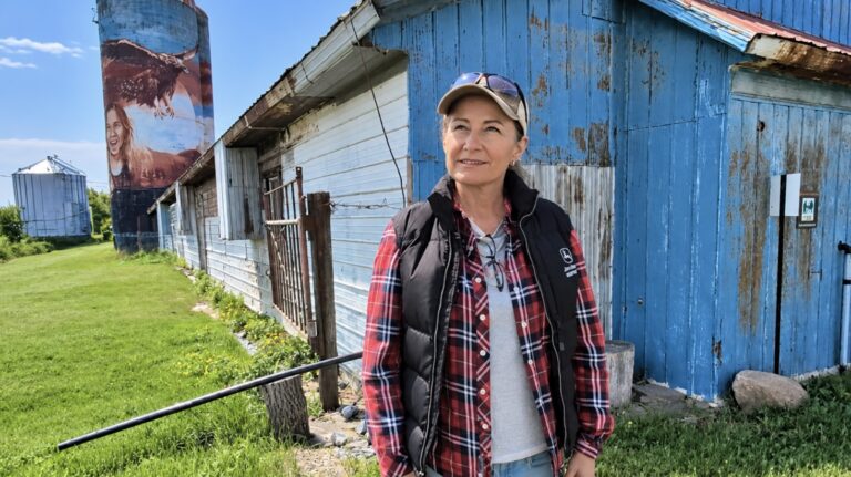 Femme debout dans une ferme devant un bâtiment agricole bleu, avec un silo peint d’une murale représentant une personne et un grand oiseau en arrière-plan.