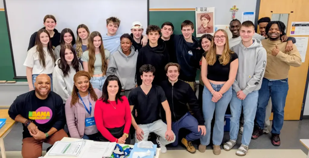 Group of about 20 youth and adults gathered in a classroom, posing together in front of a board and projection screen.