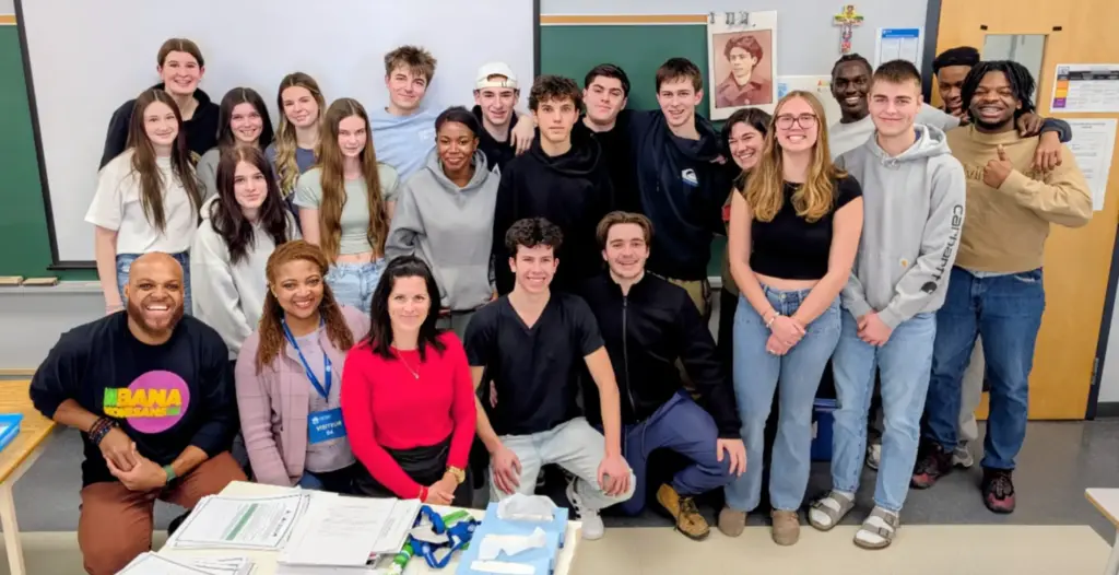 Groupe d’une vingtaine de jeunes et d’adultes réunis dans une salle de classe et posant ensemble devant un tableau et un écran de projection.