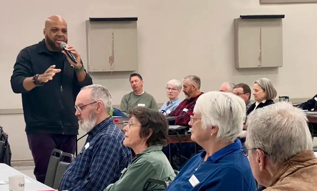 A man standing at a microphone addresses a group of adults seated around tables in a community room.