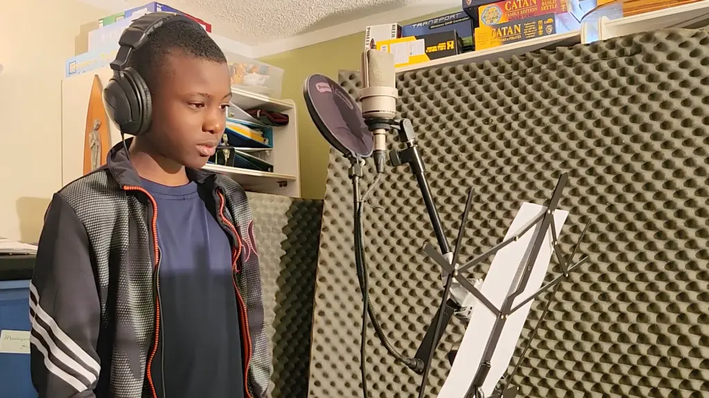 Un jeune garçon noir portant des écouteurs chante dans un microphone en studio.