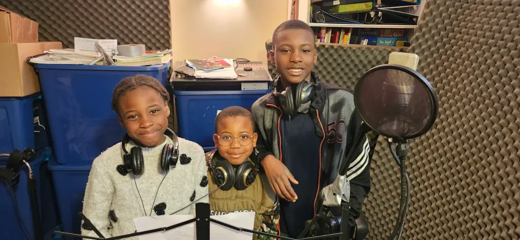 Trois jeunes enfants souriants avec des écouteurs autour du cou dans un studio.