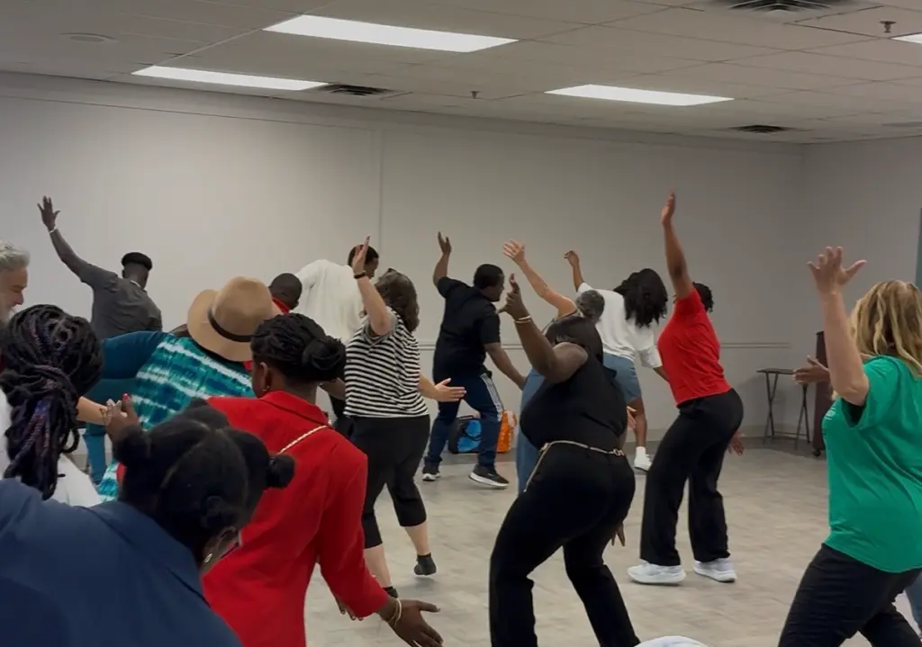 Un groupe de personnes exécute ensemble une chorégraphie dans une grande salle, plusieurs levant un bras et se penchant dans le même mouvement.