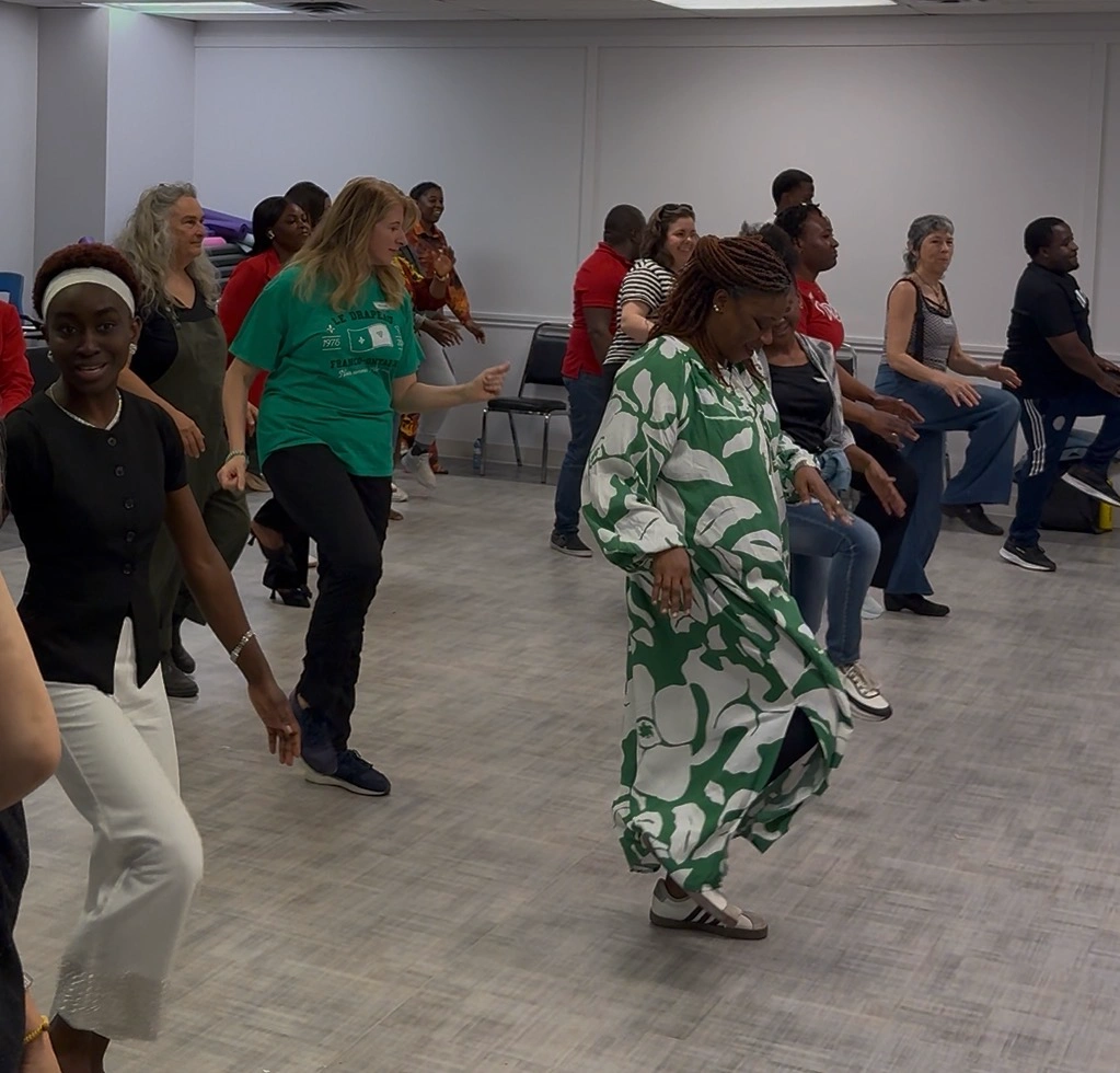 Un groupe de personnes exécute la même chorégraphie dans une grande salle, les bras abaissés et une jambe tendue ou levée dans le même mouvement.