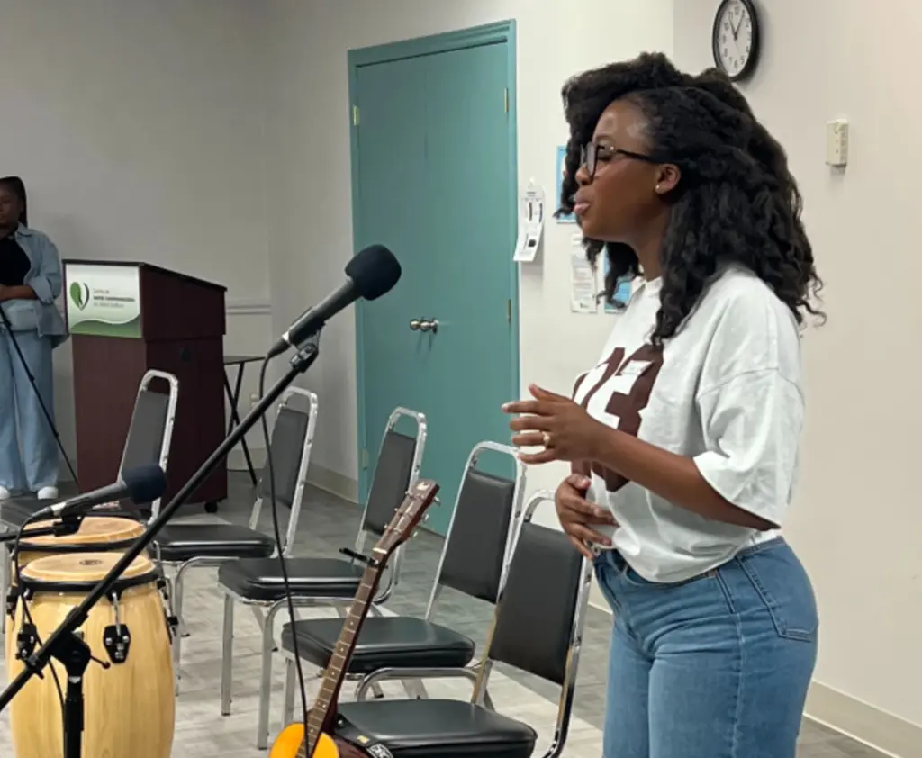 Une femme s’adresse au public devant un microphone dans une grande salle, avec une guitare et des congas à ses côtés.