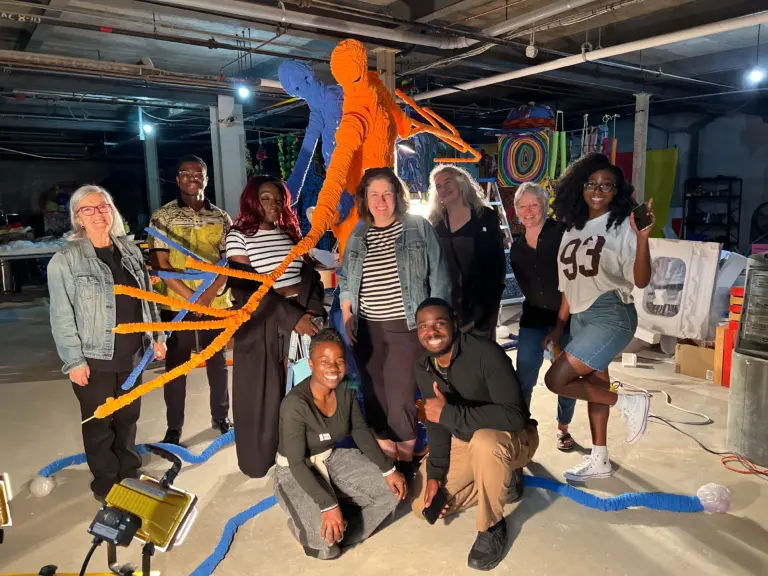 Un groupe diversifié de personnes sourit en posant devant une grande sculpture bleue et orange rappelant des figures humaines et des racines.