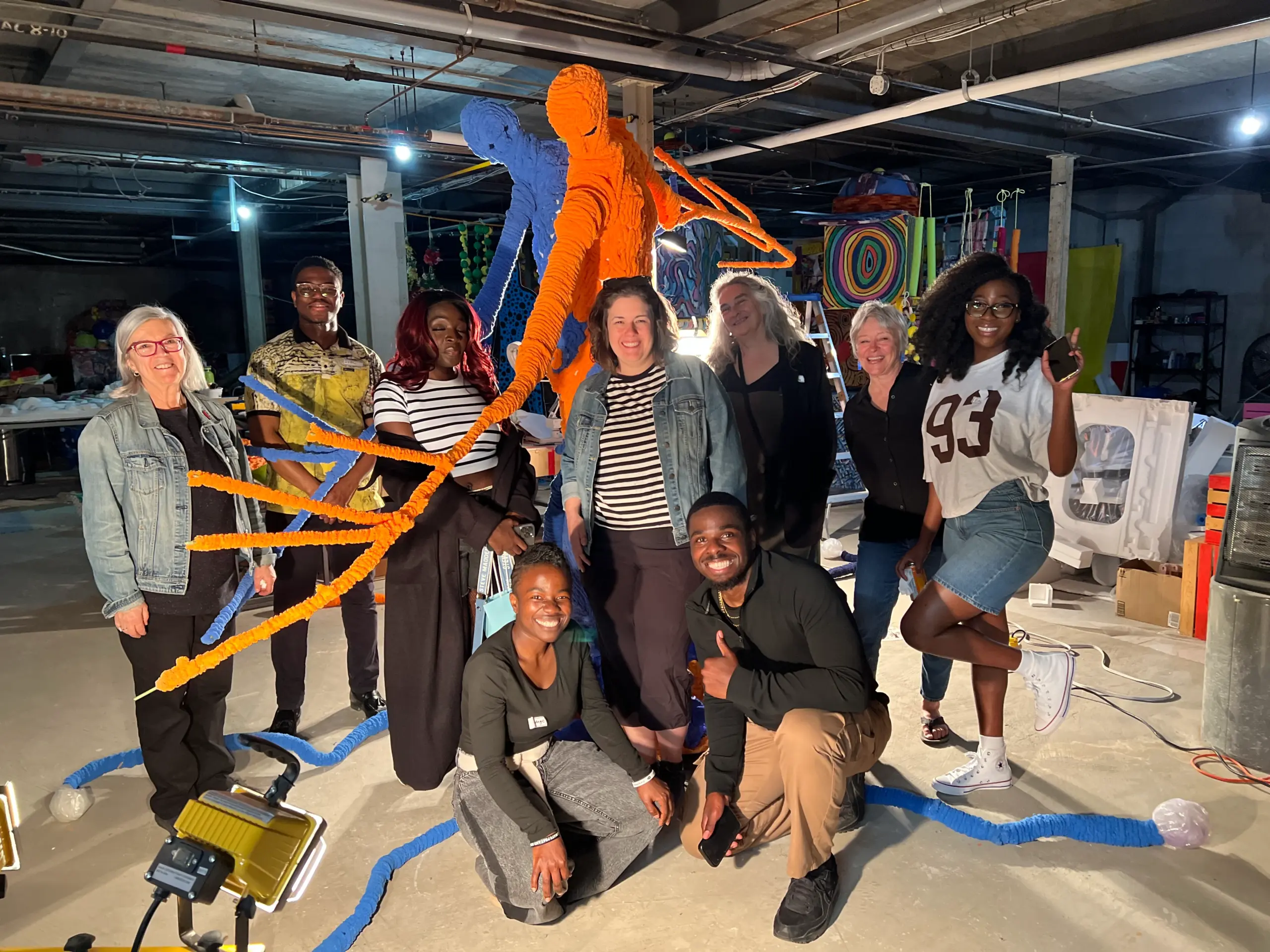 Un groupe diversifié de personnes sourit en posant devant une grande sculpture bleue et orange rappelant des figures humaines et des racines.