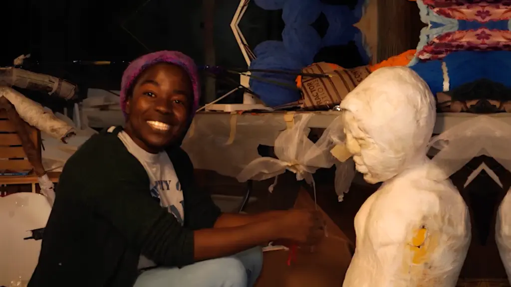 Une personne souriante portant une tuque mauve est assise dans un atelier à côté d'une sculpture blanche en forme de buste.