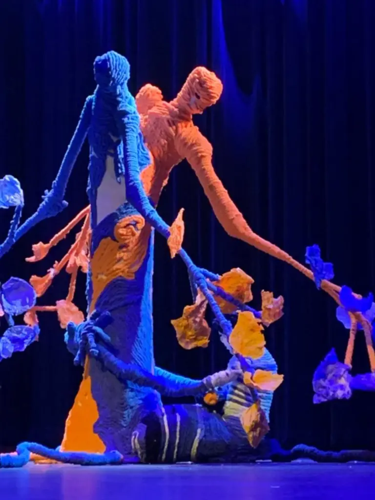 Une installation artistique composée de formes humaines et de branches recouvertes de textile bleu et orangé est présentée sur une scène éclairée en bleu.