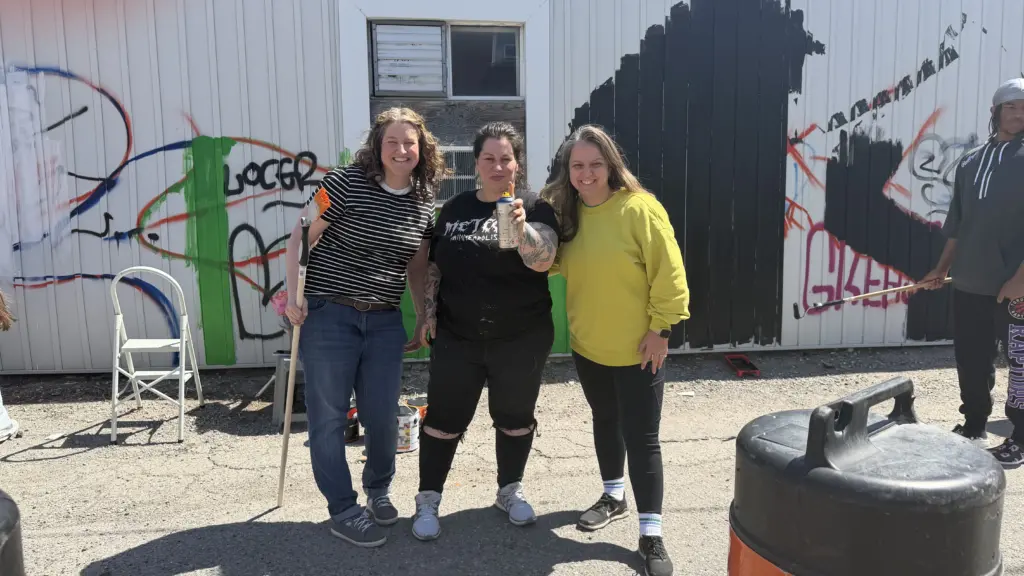 Trois femmes posent devant un mur couvert de graffitis en cours de peinture, l’une tenant un pinceau au bout d’un manche et une autre une bouteille de peinture en aérosol.
