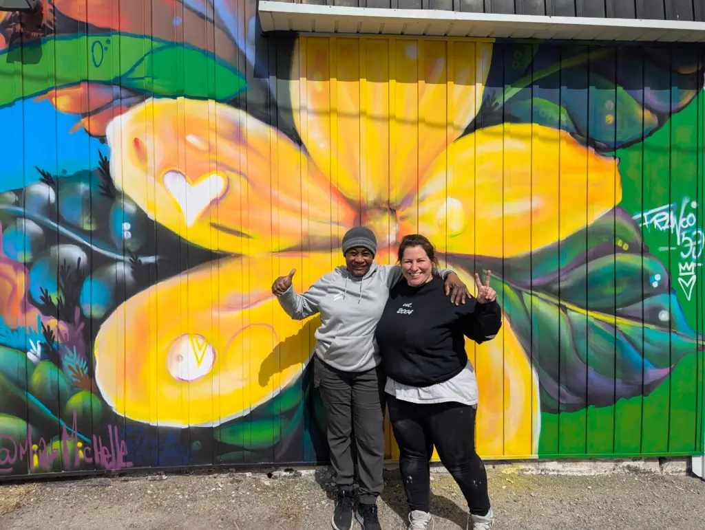 Deux personnes posent côte à côte devant une murale colorée dominée par une grande fleur jaune, l’une faisant un pouce levé et l’autre un signe de paix.