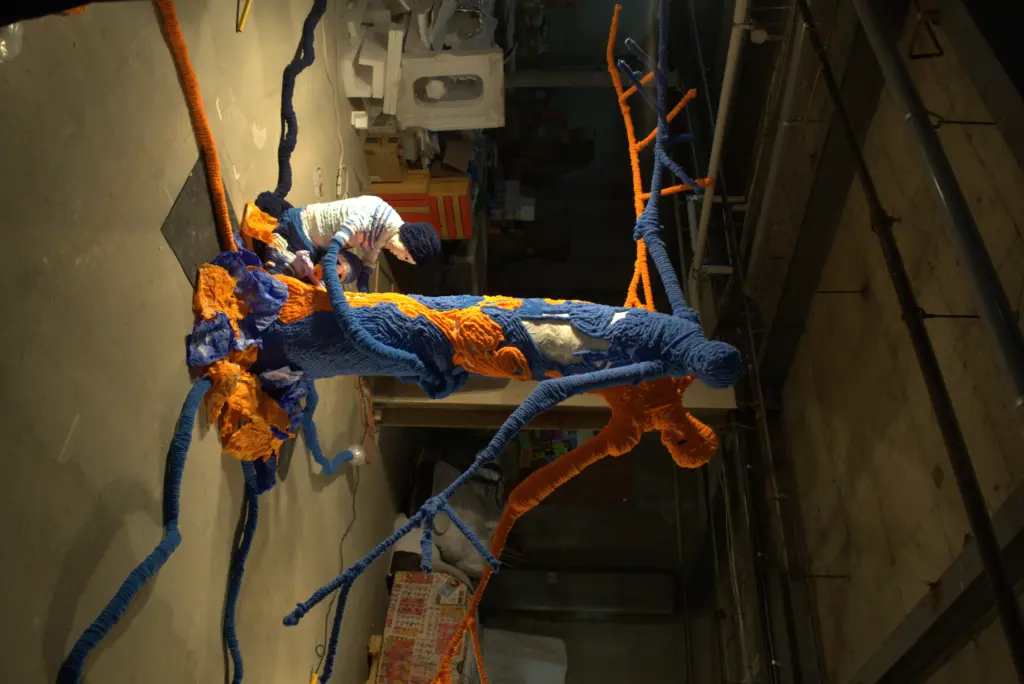An art installation made of human-like forms and branches covered in blue and orange textiles is displayed on a stage lit in blue.