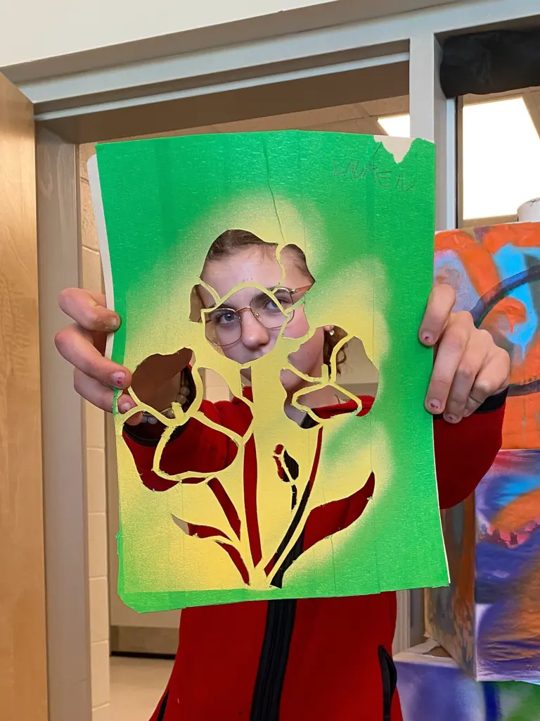Une personne tient devant son visage un grand pochoir vert et jaune découpé de fleurs et de tiges, son visage apparaissant à travers les ouvertures.