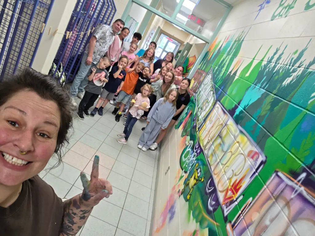 Égoportrait "selfie" de l'artiste Mique Michelle avec les élèves et le personnel devant leur nouvelle murale dans le corridor.