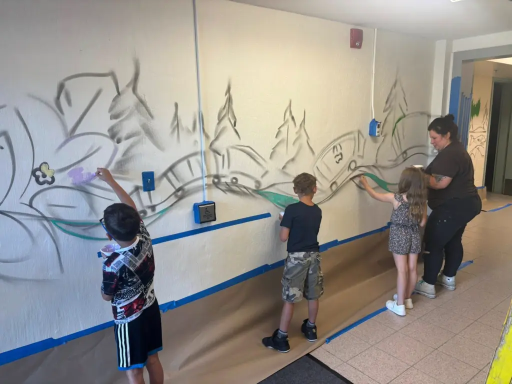 Trois jeunes élèves et l'artiste peignent en vert le bas d'un mur blanc avec les contours noirs d'une forêt et d'un train.