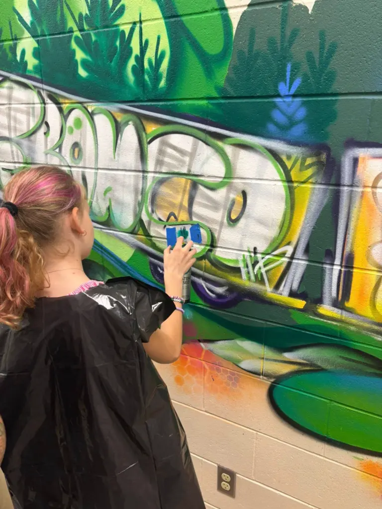 Une personne utilise une bombe de peinture et un pochoir sur une murale représentant un wagon de train, entouré de végétation verte.