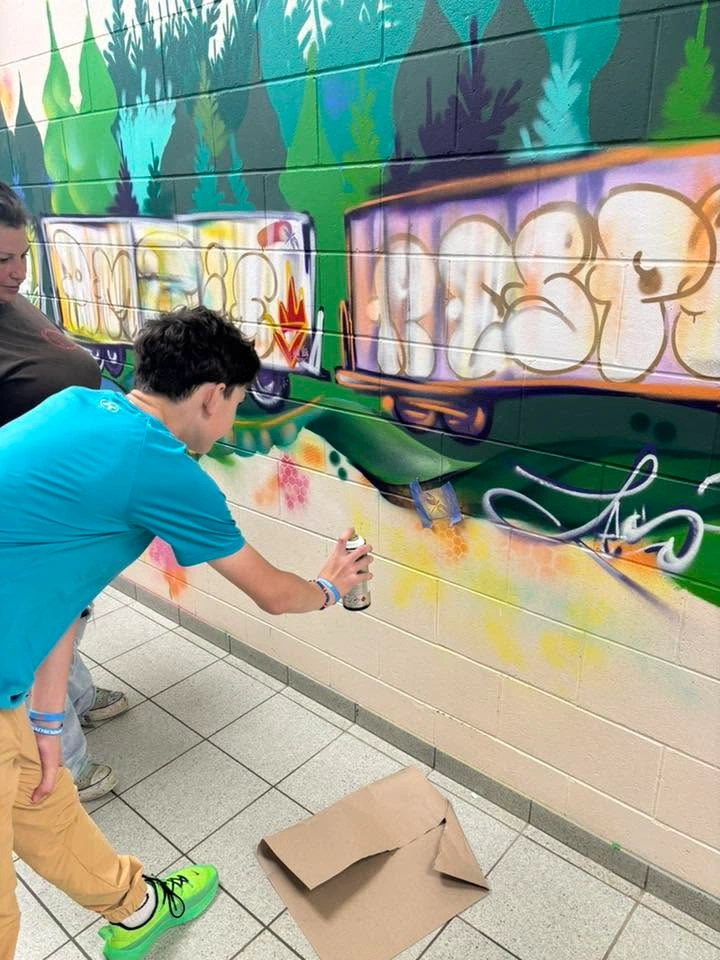 Une personne applique de la peinture en aérosol sur une murale représentant deux wagons de train entourés de végétation verte, sous le regard d’une autre personne.
