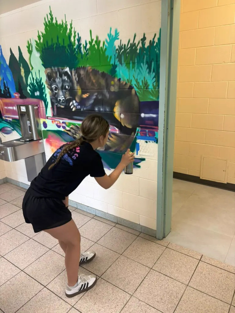 Une personne applique de la peinture en aérosol au pochoir sur une murale représentant un grand animal sombre couché devant une végétation dense.
