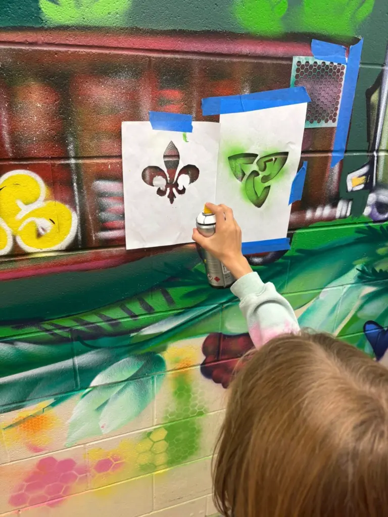 Une personne utilise une bombe de peinture et un pochoir pour ajouter des symboles de la francophonie ontarienne sur un wagon de train peint dans une murale.