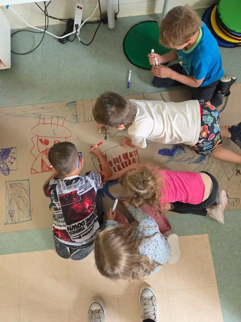 Cinq enfants dessinent ensemble avec des marqueurs sur une grande feuille de papier brun étendue au sol.