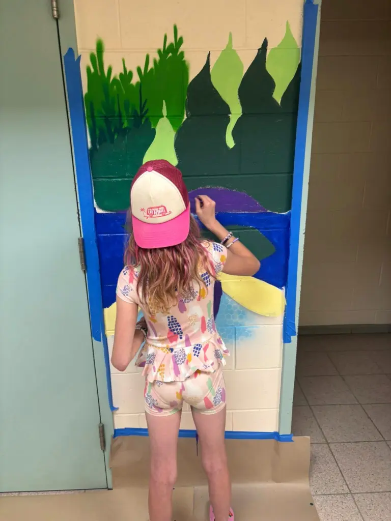 Une enfant peint une section de murale représentant un paysage composé d’eau bleue et de végétation dans différentes nuances de vert.