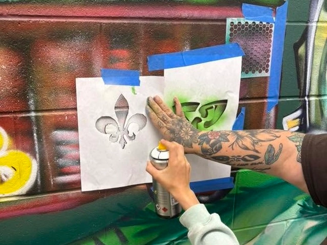 Personne utilisant une bombe de peinture et des pochoirs pour ajouter une fleur de lys et un symbole vert à une murale colorée.