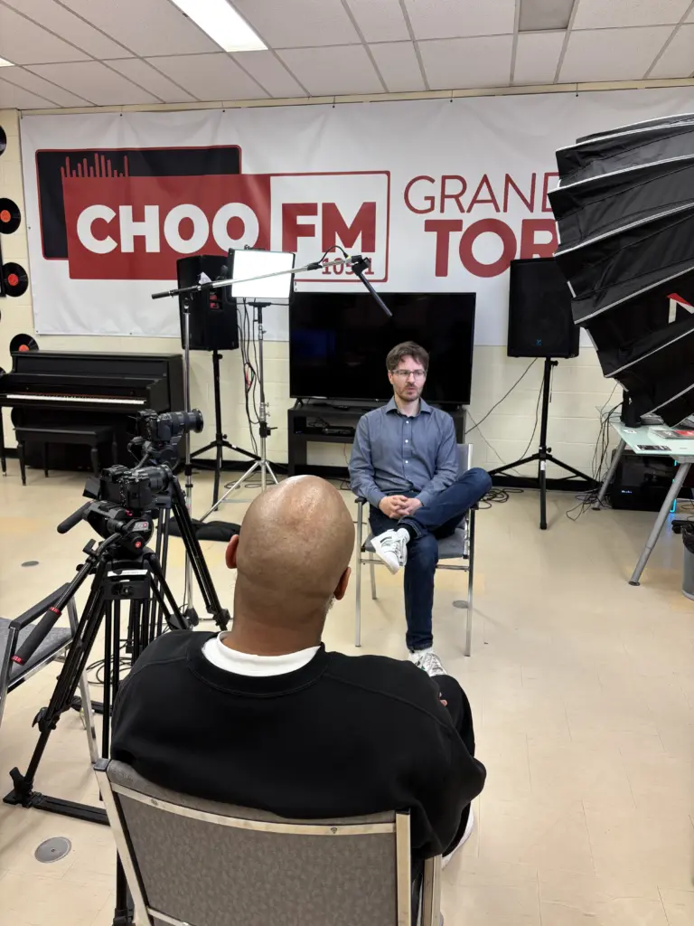Un homme est filmé par une caméra dans un studio, devant une grande bannière de la radio CHOO FM.