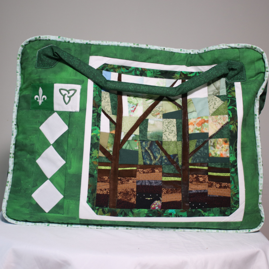 Housse de transport en tissu vert ornée d’un panneau textile représentant une forêt stylisée, avec une longue poignée et des symboles franco-ontariens sur le côté gauche.