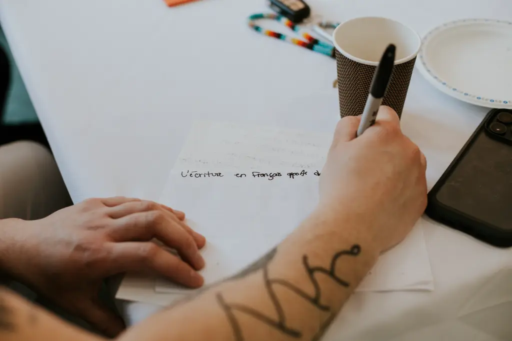 Gros plan sur la main d'une personne avec un tatouage sur le bras qui écrit un texte sur une feuille blanche avec un marqueur noir.