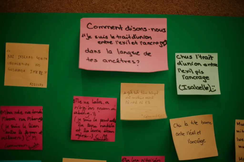 Un tableau vert couvert de feuillets adhésifs colorés traduisant la phrase « Je suis le trait d'union entre l'exil et l'ancrage » en plusieurs langues.