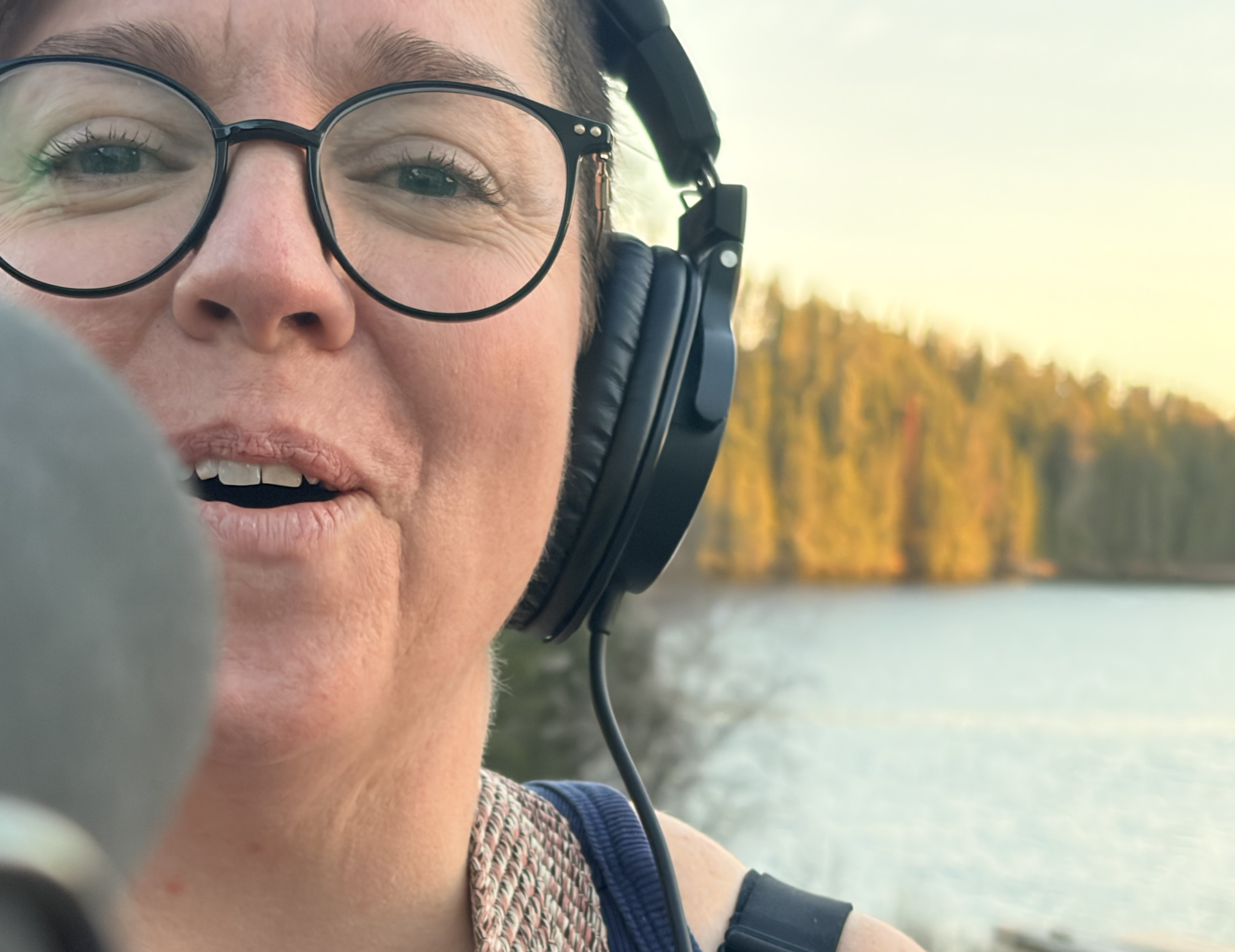 Person wearing glasses and headphones, speaking into a microphone outdoors, with a lake and wooded shoreline in the background.