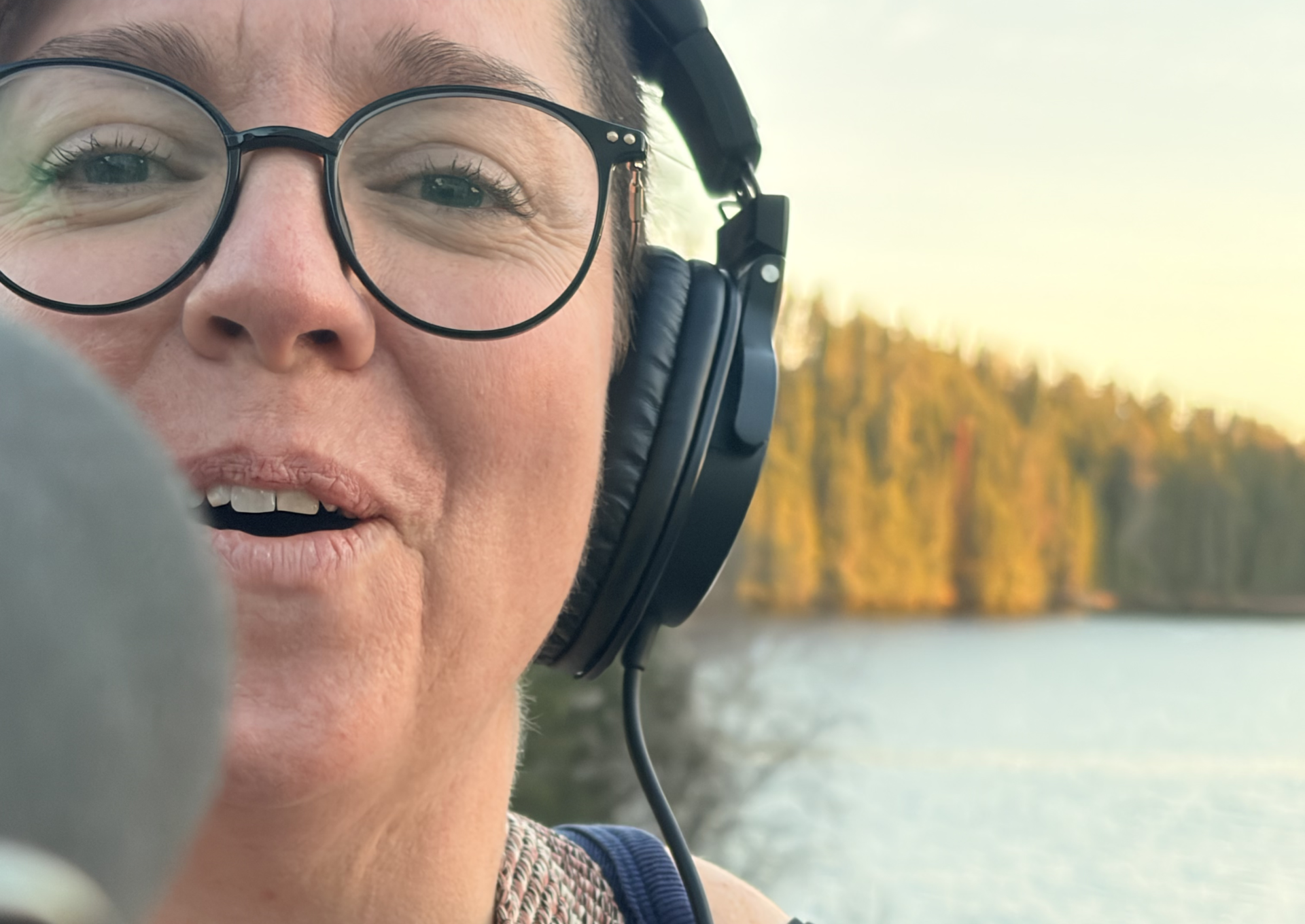 Personne portant des lunettes et un casque d’écoute, parlant devant un microphone à l’extérieur, avec un lac et une rive boisée en arrière-plan.