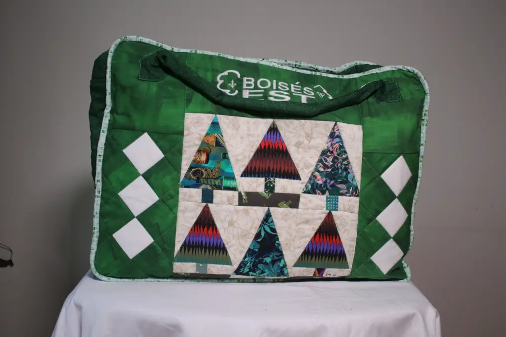 Housse de transport verte en tissu, munie d’une poignée et décorée du logo de Boisés Est, de motifs géométriques blancs et d’un panneau de courtepointe représentant des arbres stylisés.