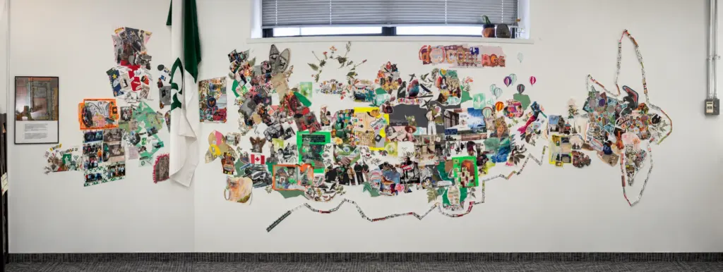 Vue panoramique d'un grand collage mural composé de nombreuses photos, coupures de presse et illustrations sur un mur blanc à côté d'un drapeau franco-ontarien.