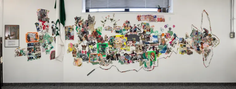 Vue panoramique d'un grand collage mural composé de nombreuses photos, coupures de presse et illustrations sur un mur blanc à côté d'un drapeau franco-ontarien.