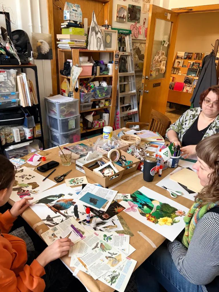 Trois personnes assises autour d'une table recouverte de papiers, d'images découpées, de ciseaux et de colle dans un atelier artistique rempli de livres.