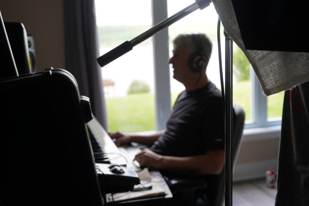 Un homme portant un casque d'écoute joue d'un clavier de studio devant une fenêtre lumineuse.