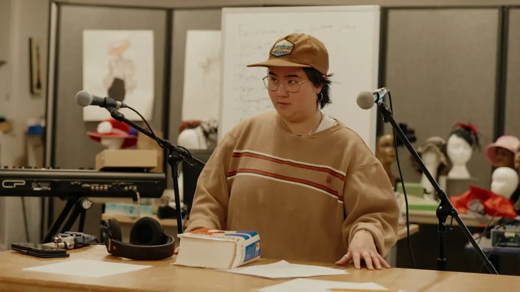 Une personne portant une casquette et un chandail brun se tient devant un microphone et du matériel audio en atelier.