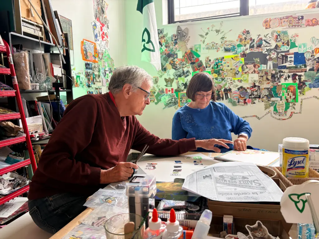 Deux personnes assises à une table travaillent à des collages, devant un mur couvert de créations et un drapeau franco-ontarien.
