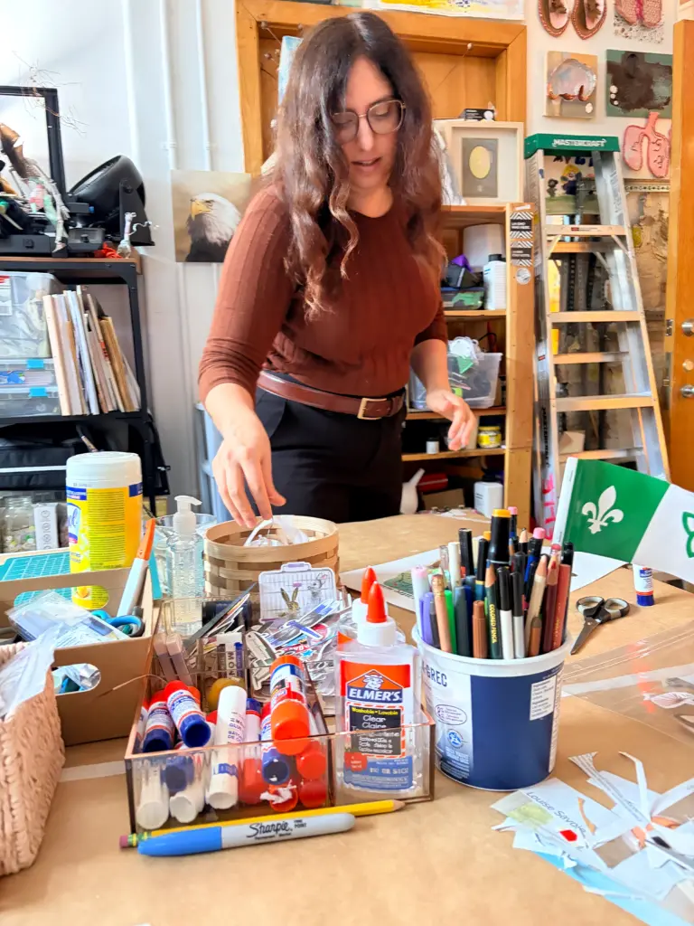 Personne préparant du matériel de création sur une table remplie de crayons, colle, ciseaux, papiers découpés et autres fournitures artistiques.