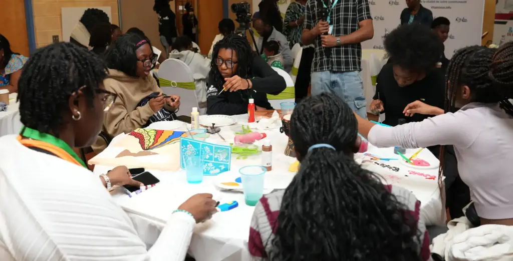 A group of young people seated around a large round table paint on canvases while being filmed by a technical crew with a camera and microphone.