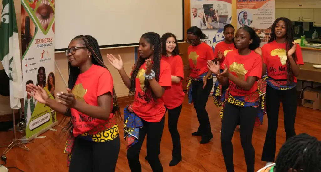 Un groupe de jeunes femmes portant des chandails rouges « Afro Nation » et des pagnes traditionnels dansent et tapent des mains devant des bannières et le drapeau franco-ontarien.