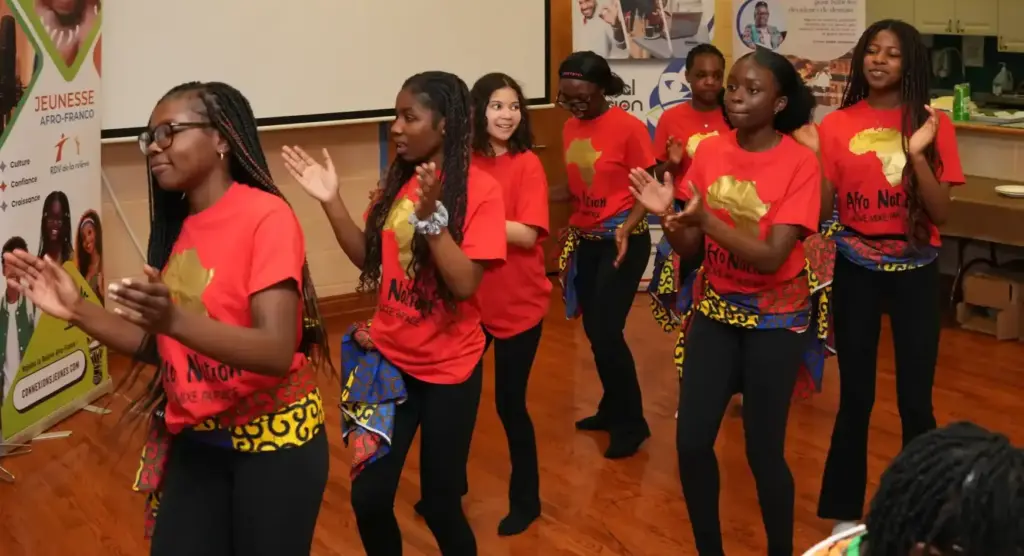 A group of young women wearing red “Afro Nation” shirts and traditional patterned wraps dance and clap in front of banners and the Franco-Ontarian flag.
