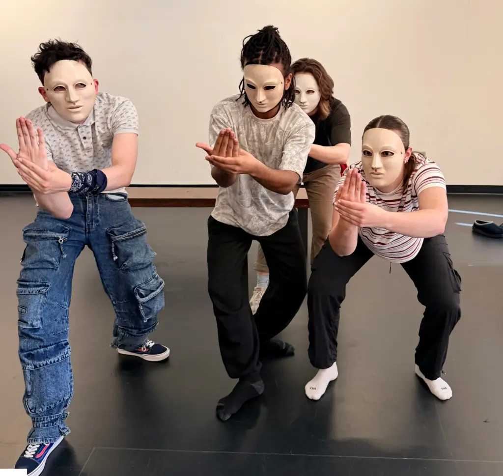 Quatre interprètes portant des masques blancs adoptent une posture chorégraphiée et synchronisée en groupe.