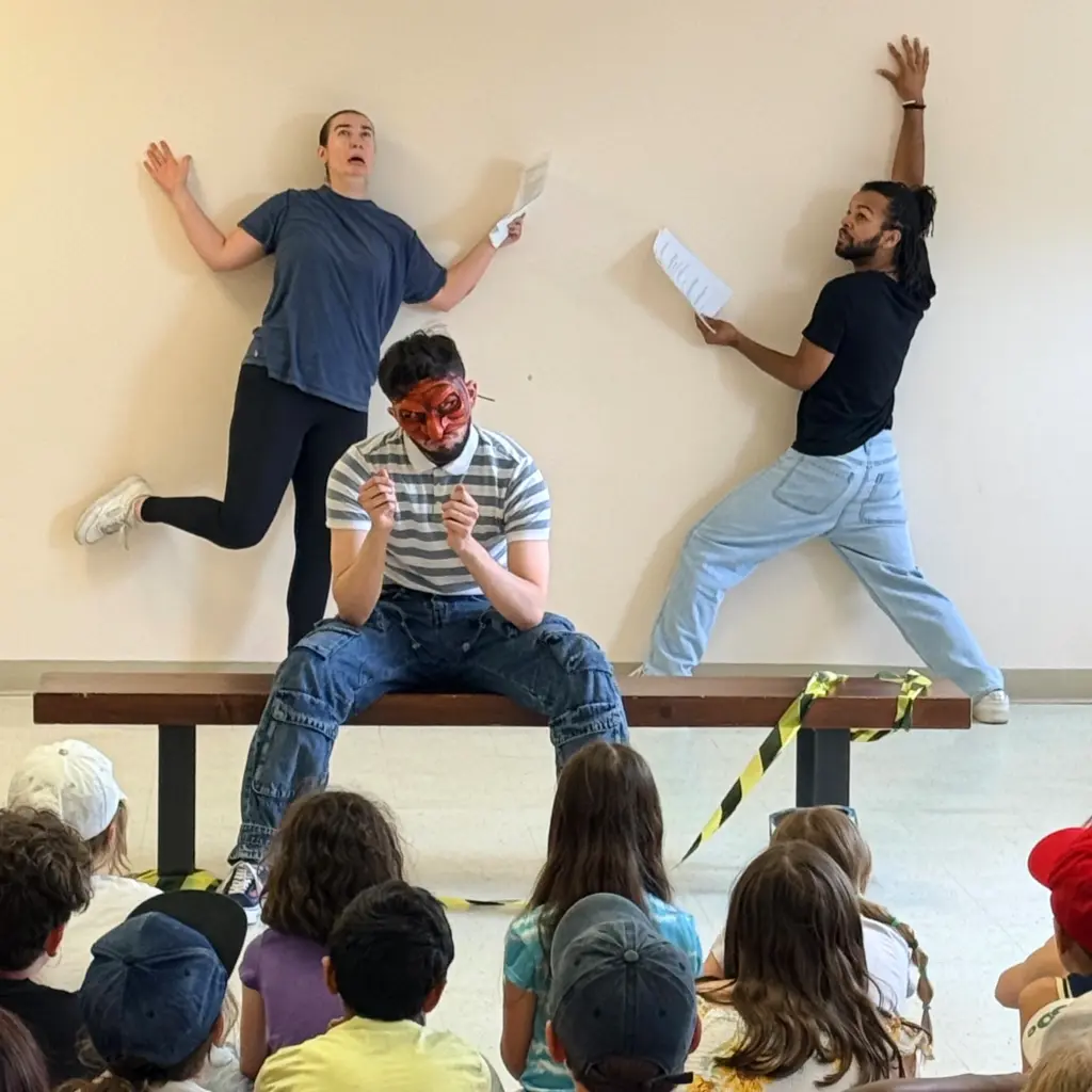 Three performers move dynamically on and around a bench in front of a wall as participants watch.
