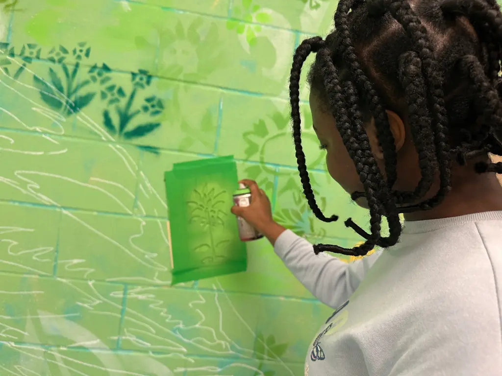 Une jeune élève utilise une bombe aérosol rose sur un pochoir pour peindre un motif floral sur le mur.