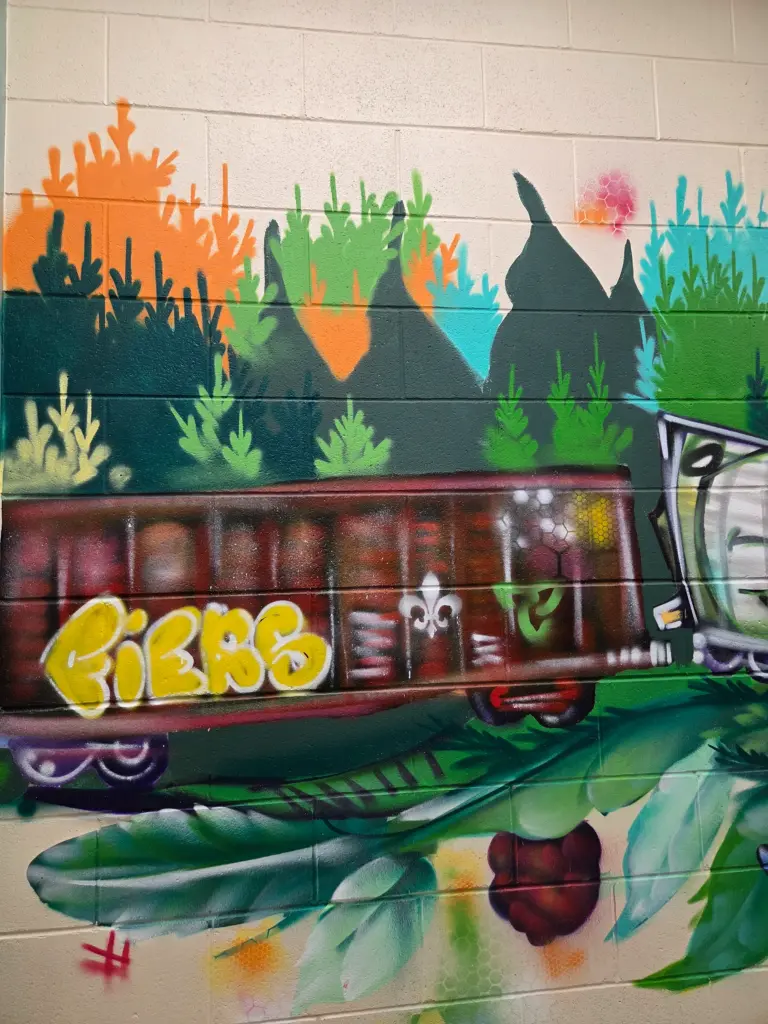 Détail d’une murale colorée représentant un wagon de train brun portant le mot « FIÈRE », entouré de végétation stylisée verte, turquoise et orange.