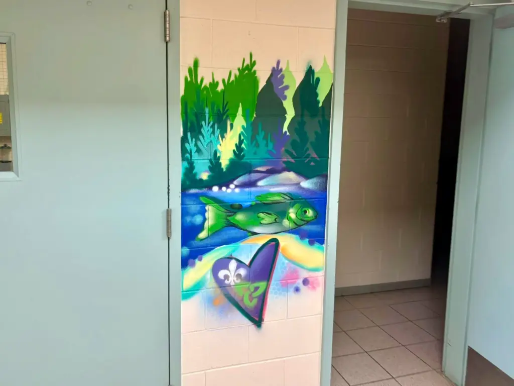 Section d’une murale intérieure représentant un poisson vert nageant dans une rivière bleue, devant une forêt stylisée, avec les symboles de la francophonie ontarienne au bas de l’image.