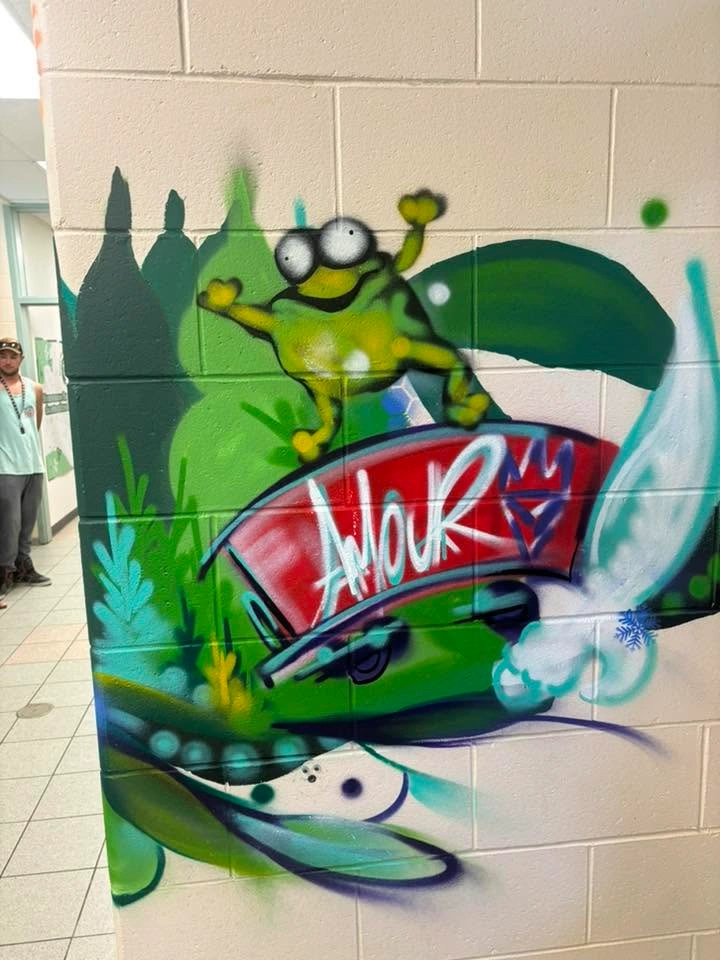 Section d’une murale intérieure montrant une grenouille verte bondissant au-dessus du mot « AMOUR », entourée de végétation verte stylisée.