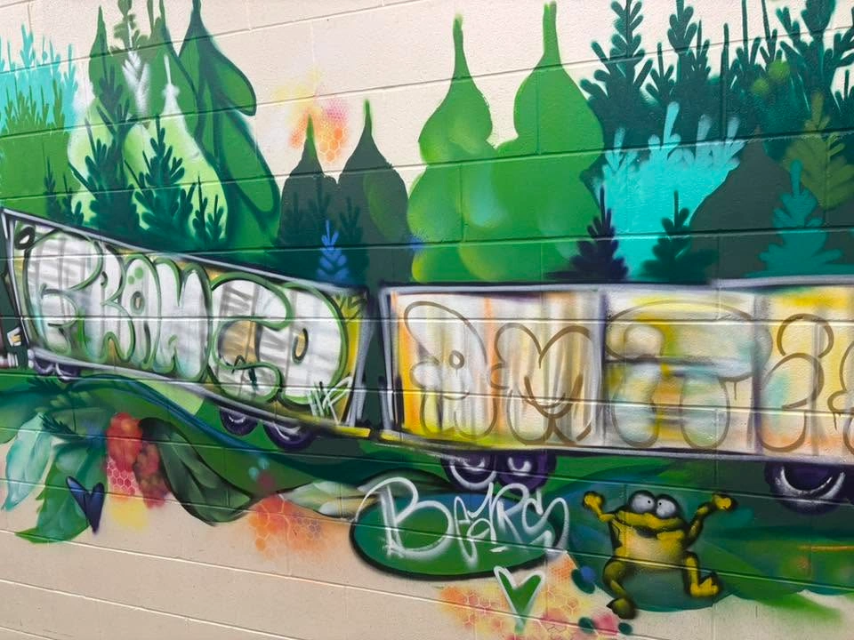 Section d’une murale intérieure montrant des wagons de train couverts de grandes lettres stylisées, entourés d’une végétation verte et turquoise, avec une petite grenouille au bas de la composition.